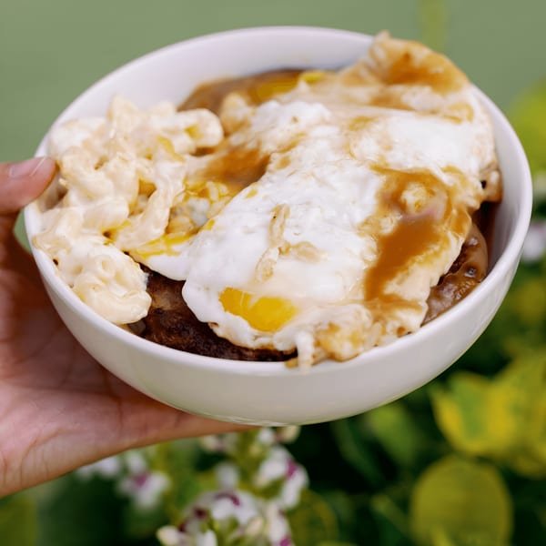 A hand holds a white bowl of L&L Hawaiian Barbecue Loco Moco with macaroni salad, eggs, and gravy.