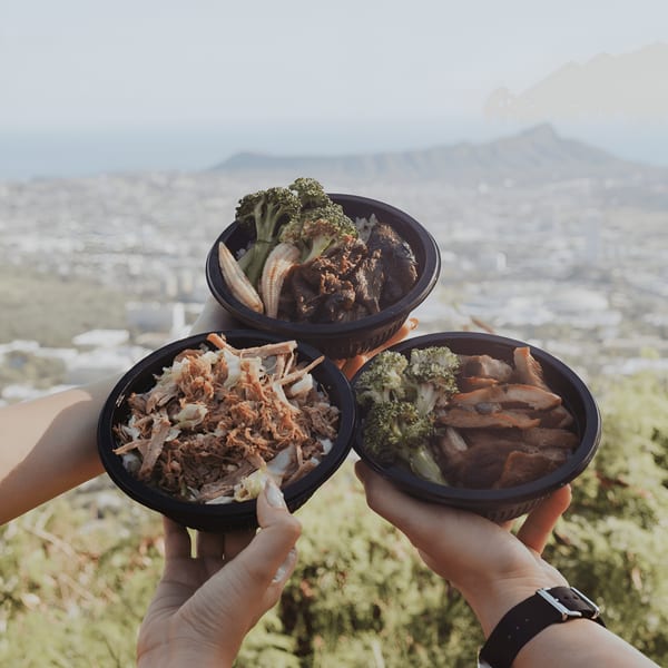 Three L&L Hawaiian Barbecue bowls with various meats and vegetables held up against a city and mountain backdrop.