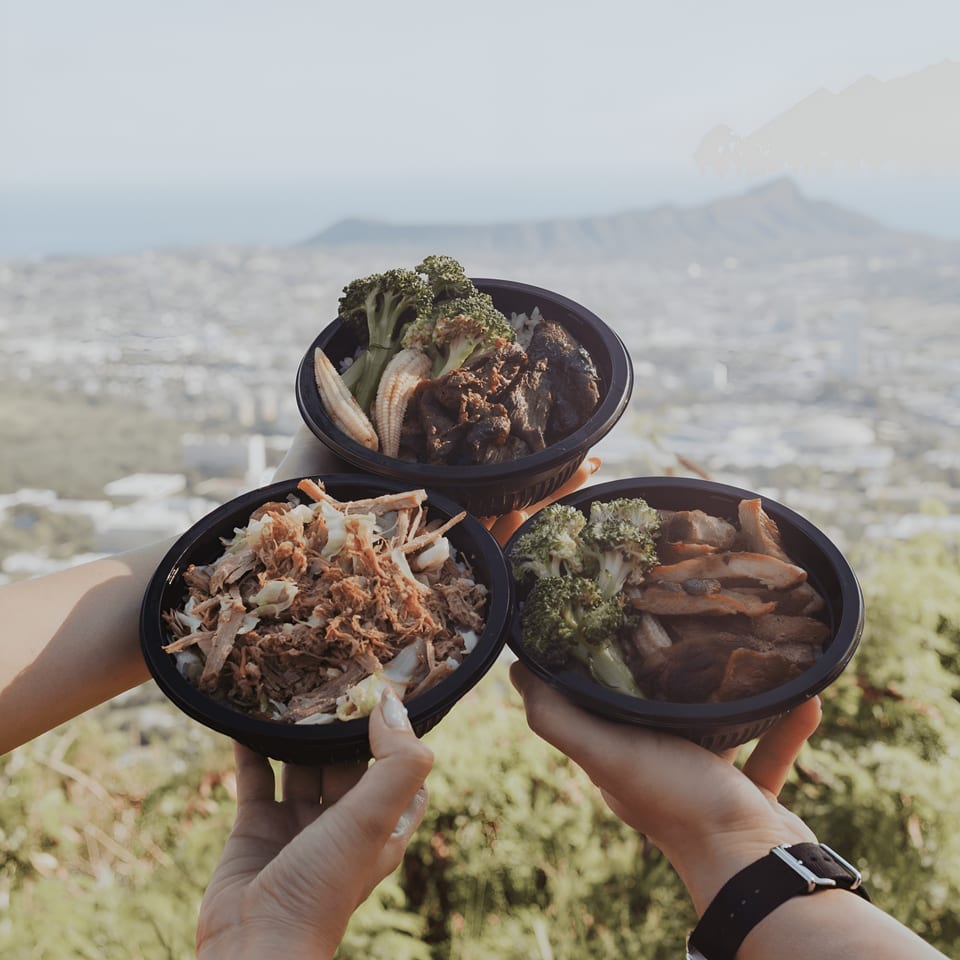 Three L&L Hawaiian Barbecue bowls with various meats and vegetables held up against a city and mountain backdrop.