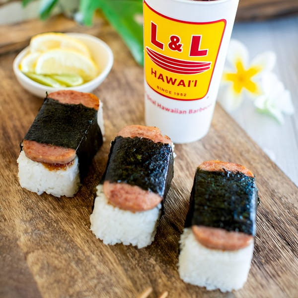 Three Spam musubi, a side of lemon slices, and an L&L Hawaiian Barbecue drink on a wooden board.