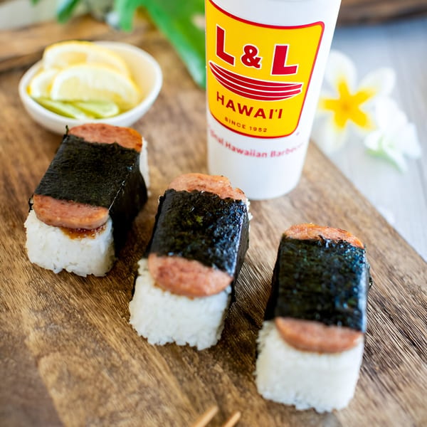 Three L&L Hawaiian Barbecue Spam Musubi on a wooden board with a drink and lemon slices.